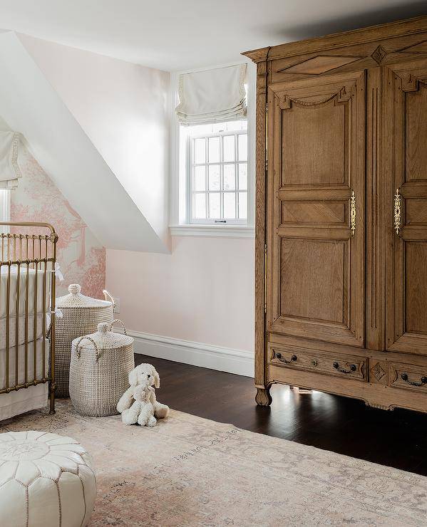 A pink French girl's nursery has a French armoire accented with antique brass hardware, a dark stained wood floor, and flanked by windows covered in off-white roman shades. I'm here. A golden metal crib sits next to a white knitted toy basket on a pink and brown rug.