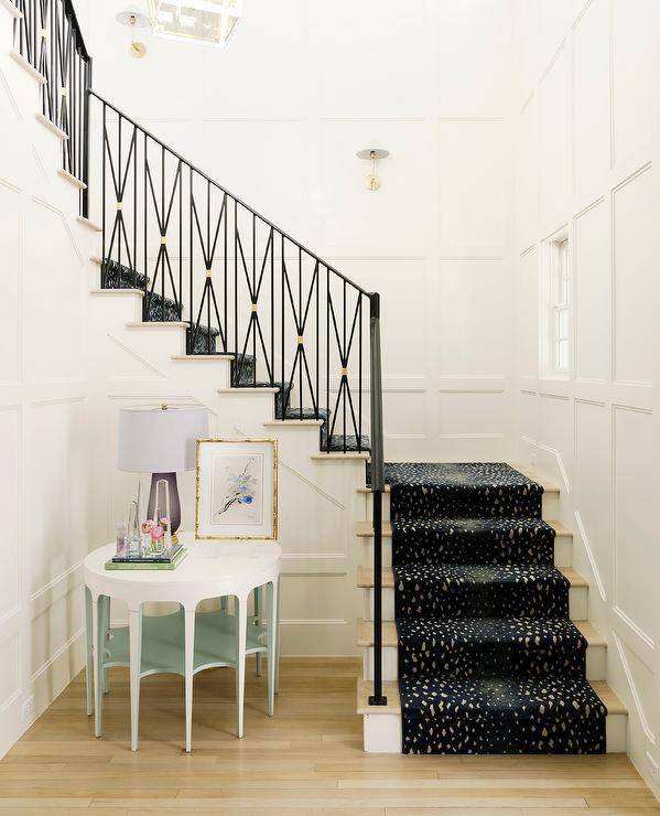 A gold and black antelope print stair runner accents a wrought iron and brass staircase and covers light wood treads fixed against white risers. A purple glass lamp sits on a round white and green table placed beside the staircase.