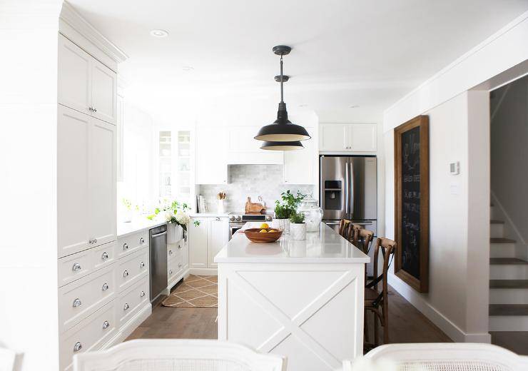 A gorgeous beveled framed chalkboard is hung in a cottage kitchen behind a white kitchen island accented with x-trim and topped with a marble alternative countertop seating French café wood and leather x-back stools. The island is lit by black barn lanterns.