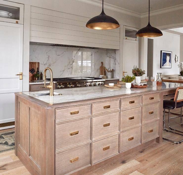 Brown apothecary center island cabinets with brass hardware are topped with a white and gold marble countertop lit by black and gold pendants and finished with a prep sink and brushed gold gooseneck faucet.