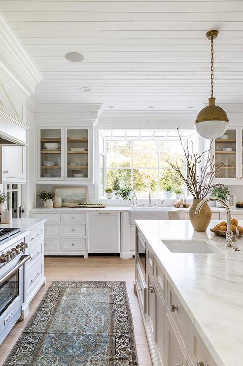 A brown and blue kitchen runner sits in a modern cottage kitchen behind a marble top kitchen island lit by brass Hicks Pendants hung over a sink with a polished nickel gooseneck faucet. Glass front cabinets flank a window located over a farmhouse sink fixed over white cabinets.