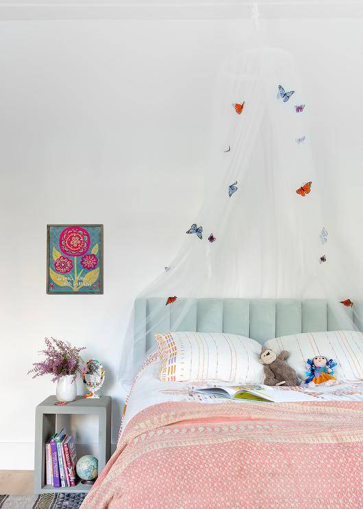 Tempat tidur berumbai saluran beludru biru muda dengan alas tidur merah muda diapit oleh meja samping tempat tidur beton berwarna abu-abu yang diakhiri dengan kanopi kupu-kupu tipis.