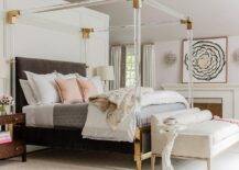 Brass and lucite canopy bed with a dark gray velvet headboard in a contemporary bedroom dressed with pink and gray bedding and accented with pink pillows and a gray quilt. Burl wood nightstand flank the bed with alabaster lamps boasting an elegant finish.