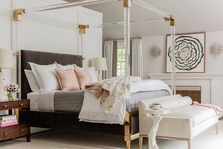 Brass and lucite canopy bed with a dark gray velvet headboard in a contemporary bedroom dressed with pink and gray bedding and accented with pink pillows and a gray quilt. Burl wood nightstand flank the bed with alabaster lamps boasting an elegant finish.