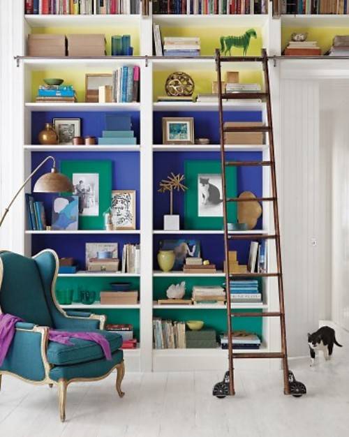 Amazing office with color blocked bookcase. Floor to ceiling built-in bookcase with levels painted different colors (Top to bottom: Martha Stewart Living Mimosa, Benjamin Moore Scandinavian Blue and Benjamin Moore Aruba Blue). Color blocked bookshelves are beautifully styled and filled with books, vases and colorful boxes. Rustic wood library ladder on white washed plank floors provide easy access to top shelves. Next to built-ins is a teal French wingback chair with purple throw and reading light is provided by vintage floor lamp.