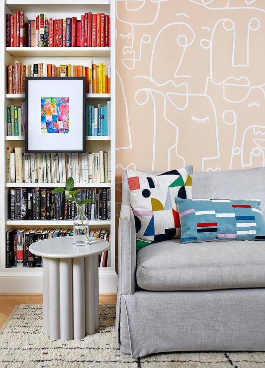 Contemporary living room features stone gray skirted sofa with round gray accent table and bookshelf with books arranged by color.