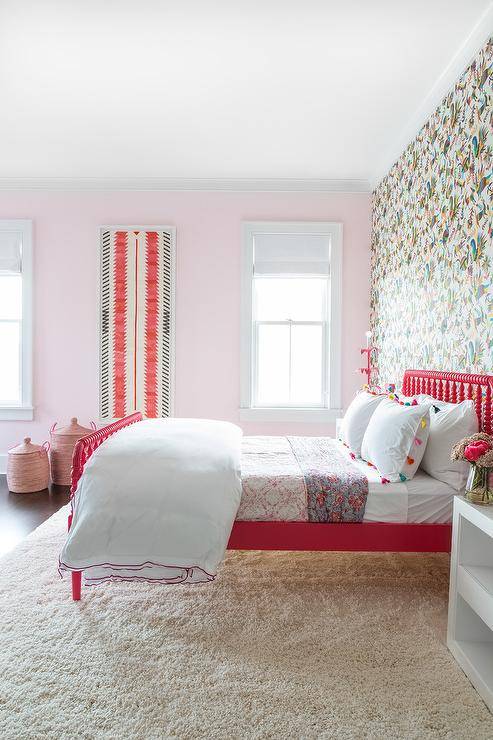 A red Jenny Lind spindle bed decorated with tassel shams and a white lacquered bedside table in the girl's bedroom. A colorful accent wallpaper gives a striking and bold look with its color and pattern balance against the soft pink adjacent wall.