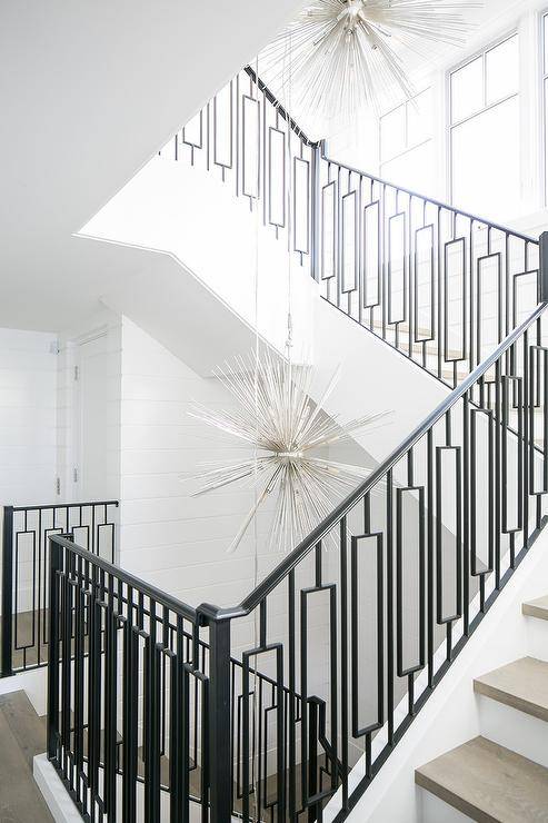 Staggered silver sputnik chandeliers illuminate a staircase fitted with gray treads and wrought iron spindles with a wrought iron railing contrasting white shiplap walls.