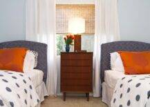 Blue and orange boy's bedroom with two twin sized beds flank either side of a mid-century modern nightstand with modern wooden table lamp. The nightstand sits in front of a bright window dressed with a woven blind and white cotton drapes. Denim blue colored headboards are paired with crisp white sheets, burnt orange pillows and white duvets with blue polka dots.