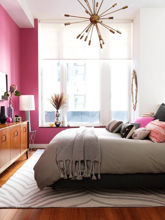 Fabulous pink and gray bedroom with bright pink accent walls and large windows with light diffusing window blinds. The bedroom features a gray upholstered bed layered with an array of pink, black and gray pillows over a gray duvet. A chunky gray knit throw drapes over the foot of the bed. A Mid-Century style dresser stands against the wall at the foot of the bed below a small flat screen tv. In the corner of the bedroom stands a small modern accent table topped with a tall gray lamp. Hardwood floors are layered with a West Elm Safari Rug that anchors the bed. A DWR Satellite Chandelier adds interest to the tall ceilings.