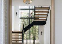 A floating staircase boasts a gold and black railing and is hit by natural light streaming in front tall windows.