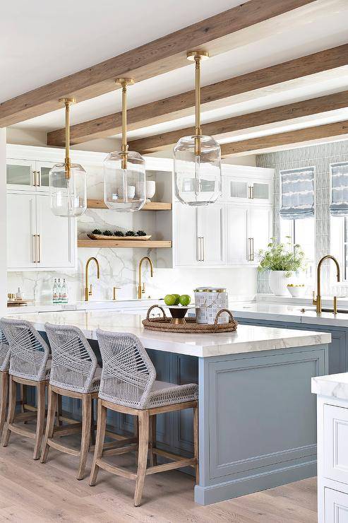 Dual blue shaker kitchen island complement a white kitchen. Gray rope stools sit at one island lit by three brass and glass lanterns hung from a stained wood beam. The other island is finished with a sink and a brass gooseneck faucet.