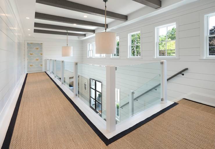 Second floor landing features gray beams on ceiling dotted with pot lighting and linen drum light pendants over a clear glass stair railing and staircase lined in a black bound sisal runner.