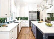 White kitchen cabinets hold a stainless steel sink with a polished nickel gooseneck faucet mounted to a marble-look countertop under a window flanked by white upper cabinets and partially framed by green hexagon backsplash tiles. Two Suzanne Kasler Morris Lanterns illuminate a blue island.