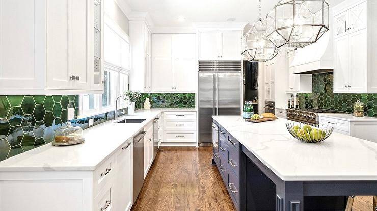 White kitchen cabinets hold a stainless steel sink with a polished nickel gooseneck faucet mounted to a marble-look countertop under a window flanked by white upper cabinets and partially framed by green hexagon backsplash tiles. Two Suzanne Kasler Morris Lanterns illuminate a blue island.