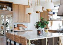 A brown wood island with brass legs is accented with a striped marble countertop seating four brown woven stools facing a sink wiht a polished nickel gooseneck faucet lit by three white and gold pendants.