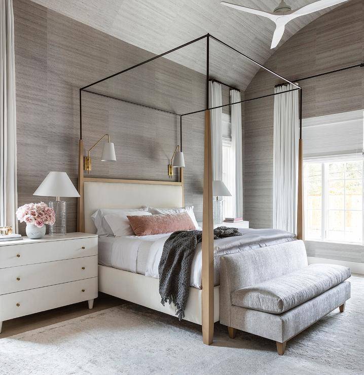 Bedroom features an ivory and brown canopy bed with pink velvet lumbar pillow lit by sconces under a barrel ceiling with a ceilng fan, a gray velvet settee at the foot of the bed, white dresser nightstand lit by fluted glass lamps and gray grasscloth wallpaper.