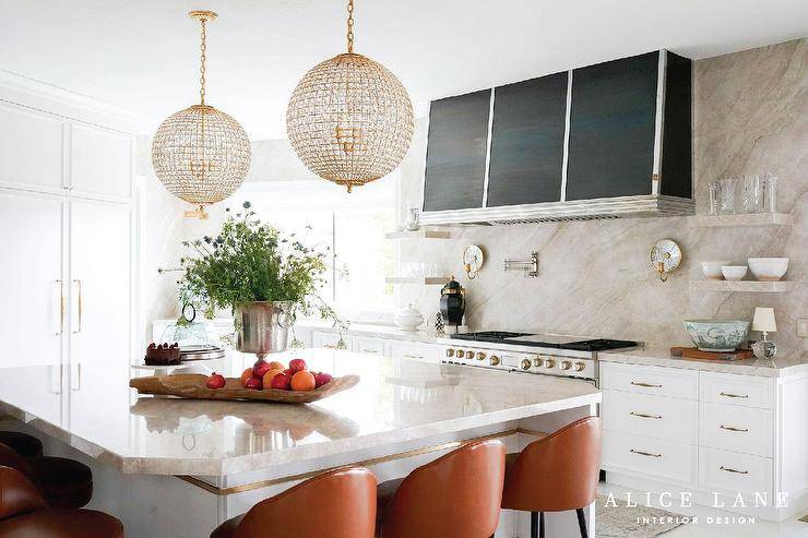 Orange leather stools sit around a white kitchen island topped with a taupe marble countertop and lit by glam crystal chandeliers. A black and silver hood is fixed between stacked taupe marble floating shelves fixed against a taupe marble backsplash. A polished nickel swing arm pot filler is mounted above a Wolf dual range flanked by white shelves with brass pulls.
