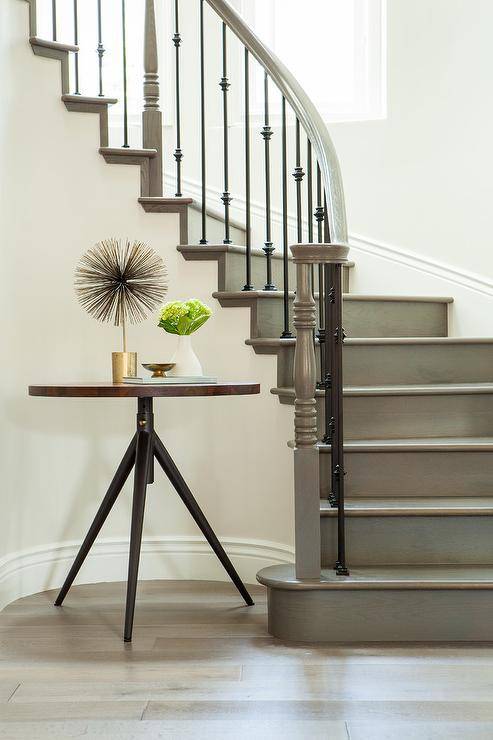 Curved gray wood staircase offers curvature and texture accented with iron spindles in a traditional foyer. Gray stair treads keep a neutral appeal merging with blond oak wood floors displaying a wood and metal round foyer table.