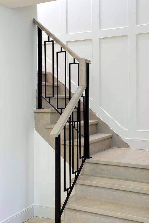 A gray wash wood staircase is finished with hollow iron balusters alongside a staircase wall clad in wainscoting.