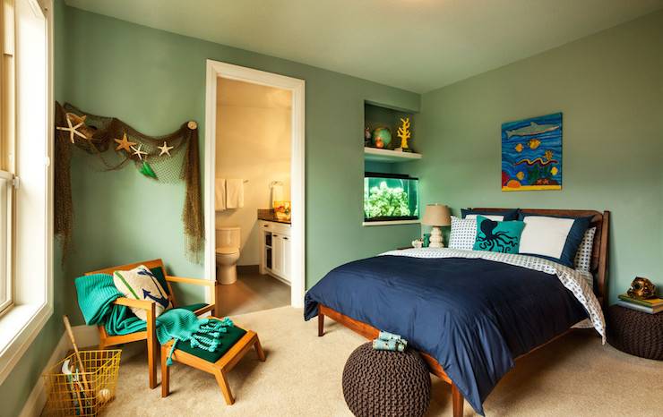 Nautical boy's room with green walls, painted Sherwin Williams Interesting Aqua, framing alcove next to bed filled with a small aquarium below a floating shelf. Kid's ocean art hangs over the wooden bed dressed with white and navy bedding accented with a turquoise and navy octopus pillow with a pair of brown knitted poufs on the floor. A Mid-Century style chair with ottoman stands across from the bed layered with a jade green throw and anchor pillow below swagged netting studded with starfish.