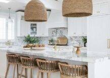Wood and leather strap stools are placed at a white plank island topped with a marble countertop holding a prep sink beneath a brass gooseneck faucet kit lit by woven basket chandeliers and natural light from skylights.