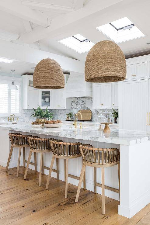 Wood and leather strap stools are placed at a white plank island topped with a marble countertop holding a prep sink beneath a brass gooseneck faucet kit lit by woven basket chandeliers and natural light from skylights.