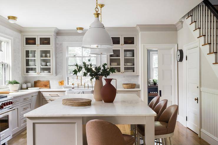 Brown leather stools sit at a honed marble countertop accenting a light gray island lit by white and gold industrial pendants hung in a light gray kitchen.