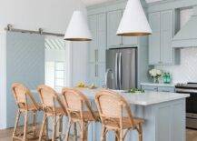 Serena and Lily Sunwashed Riviera Counter Stools sit at a gray blue kitchen island illuminated by Goodman Hanging Lamps hung on either side of an island sink. A stainless steel refrigerator is recessed under gray blue cabinets.