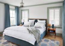 Chic blue bedroom features off-white walls accented with gray crown moldings and baseboards lined with a blue velvet bed with wood frame dressed in white and blue bedding as well as a gray throw blanket flanked by wood mid-century modern nightstands and blue lamps.