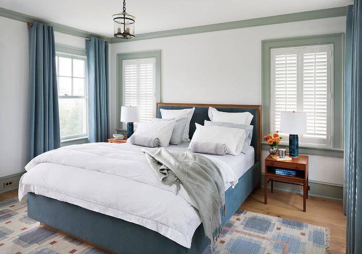 Chic blue bedroom features off-white walls accented with gray crown moldings and baseboards lined with a blue velvet bed with wood frame dressed in white and blue bedding as well as a gray throw blanket flanked by wood mid-century modern nightstands and blue lamps.