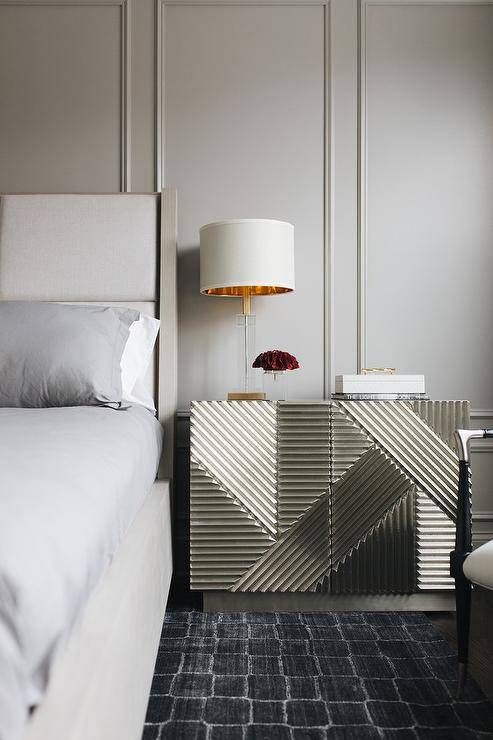 A modern gold cabinet topped with a glass and gold lamp, sits on a black rug in front of a gray wainscoted wall and beside a light gray wingback bed dressed in platinum gray bedding.