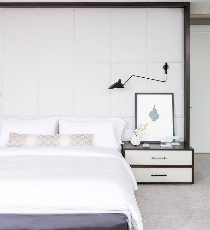 A full-wall white padded headboard accents a king-sized bed and is fitted with a black swing arm sconce lighting white bedding complemented with a taupe lattice lumbar pillow. An abstract art piece sits on a white and brown nightstand beside the bed.