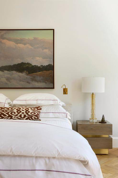 Modern bedroom features a low gray headboard with purple hotel bedding under art and a wood and brass nightstand lit by a brass lamp.