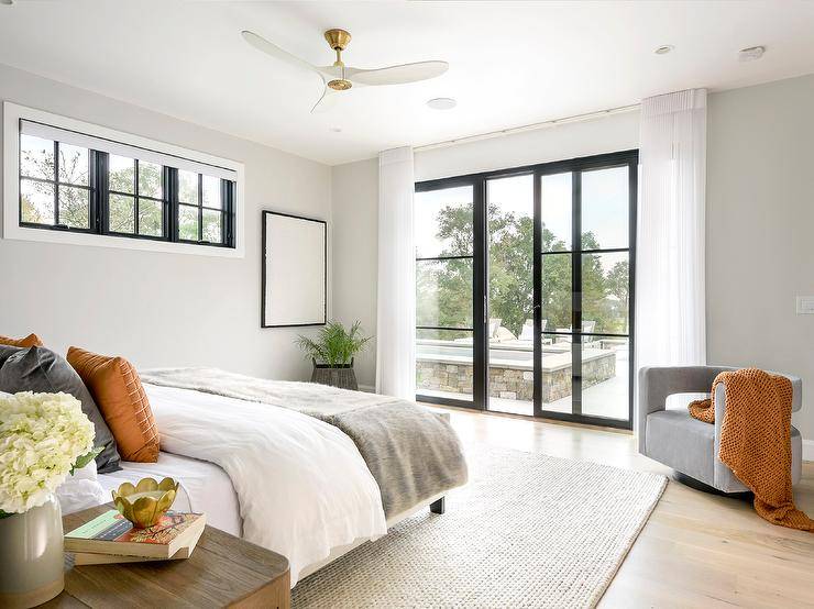 Spacious bedroom features a black platform bed accented with orange and gray velvet pillows atop a cream rug, a gray accent chair, black framed windows and a white and gold modern ceiling fan.