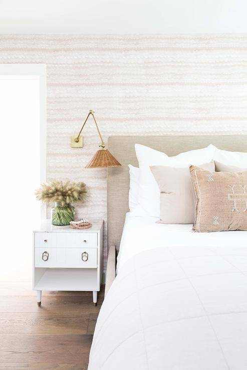 Beige fabric upholstered bed with a marble top nightstand in a transitional bedroom designed with a rattan and brass swing arm sconce on a pink and white wallpaper wall.