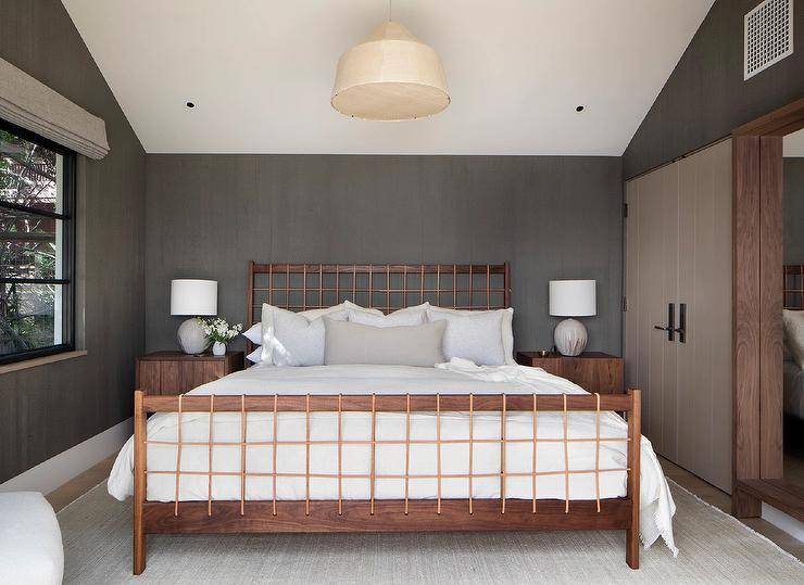 Brown wooden nightstands accented with concrete ball lamps sit on either side of a wooden bed with leather straps. The bed sits on a light gray rug in front of a gray wallpapered wall.