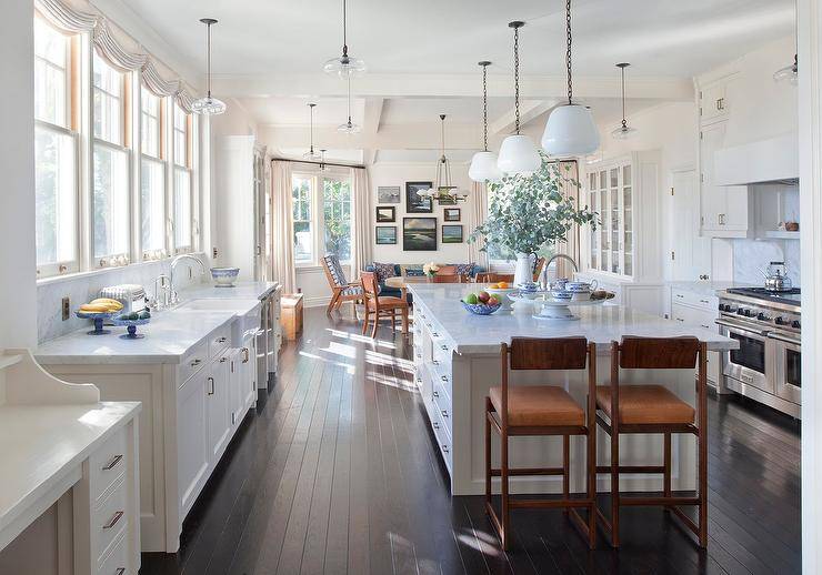 Milk glass lanterns light a marble top island finished with a sink and paired with brown wood and leather chairs placed on a dark oak thin plank floor. A row of windows is located over an apron sink with a chrome deck mount faucet fixed to a marble countertop over white cabinets.