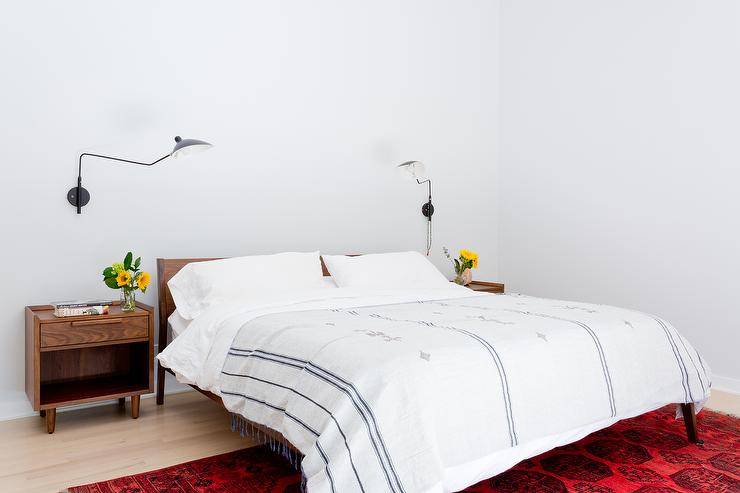 Brown oak nightstands lit by oil rubbed bronze swing arm sconces sit flanking a brown oak mid-century modern bed dressed in white and blue French linen bedding and placed on a red vintage rug.