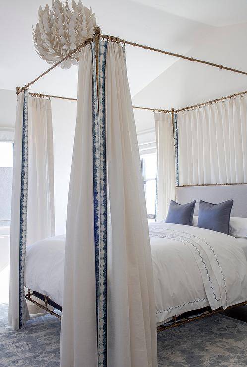 Tirai putih dan biru melengkapi tempat tidur kanopi kuningan dengan headboard ungu ungu yang ditempatkan di belakang bantal beludru ungu yang ditempatkan di atas tempat tidur putih dengan hiasan bergigi ungu.