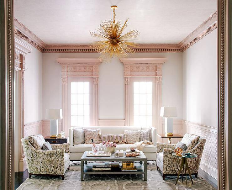 Pink crown moldings accented with dentil trim complement light gray living room walls and windows framed with pink moldings. Beneath the windows, a white shelter back sofa sits on a gray rug facing a gray coffee table positioned between facing pink and gray accent chairs. The room is illuminated by a brass sputnik light.