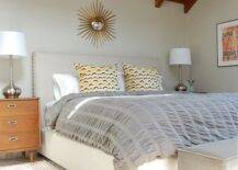 Attic bedroom with gold sunburst mirror over upholstered bed with nailhead trim and gray pintuck comforter with fun retro yellow pillows. Gray bench at foot of bed and vintage nightstands with silver lamps. Bedroom with gray wall color and West Elm Jute Chenille Herringbone Rug.