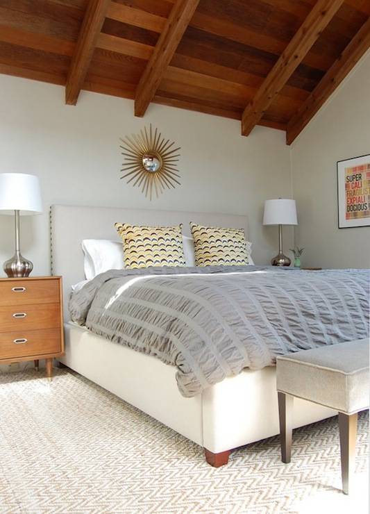 Attic bedroom with gold sunburst mirror over upholstered bed with nailhead trim and gray pintuck comforter with fun retro yellow pillows. Gray bench at foot of bed and vintage nightstands with silver lamps. Bedroom with gray wall color and West Elm Jute Chenille Herringbone Rug.