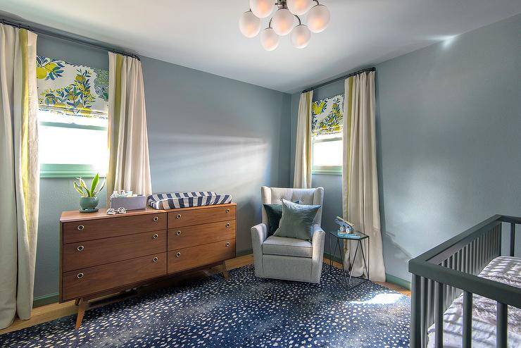 Blue boys nursery features a large blue antelope pattern area rug, a lemon print Roman shade, and a mid-century modern dresser. A gray tufted wingback glider is designed in the corner of the room between two windows with cream and yellow curtains.