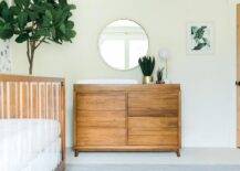 A round silver mirror hangs over a brown vintage dresser topped with a white changing pad. The dresser sits between a hung green abstract art piece and a fiddle leaf fig plant.