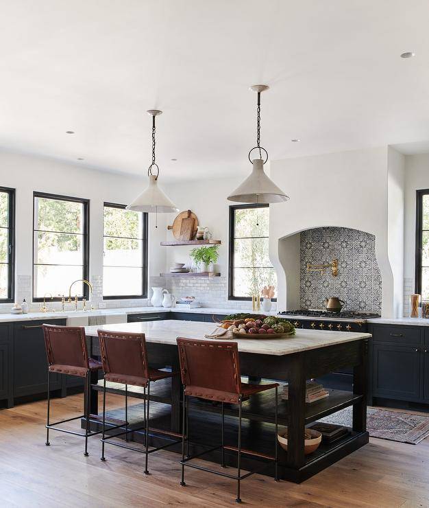 A dark brown open island seats brown leather stools at a honed white marble countertop lit by two vintage lanterns in a stunning brown and blue Mediterranean style kitchen.