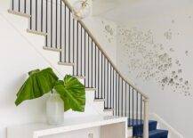 Eclectic foyer features a light taupe staircase lined with a royal blue velvet stair runner as well as butterfly wall art alongside a pair of wine jugs tucked under a white waterfall console table with overlay trim.