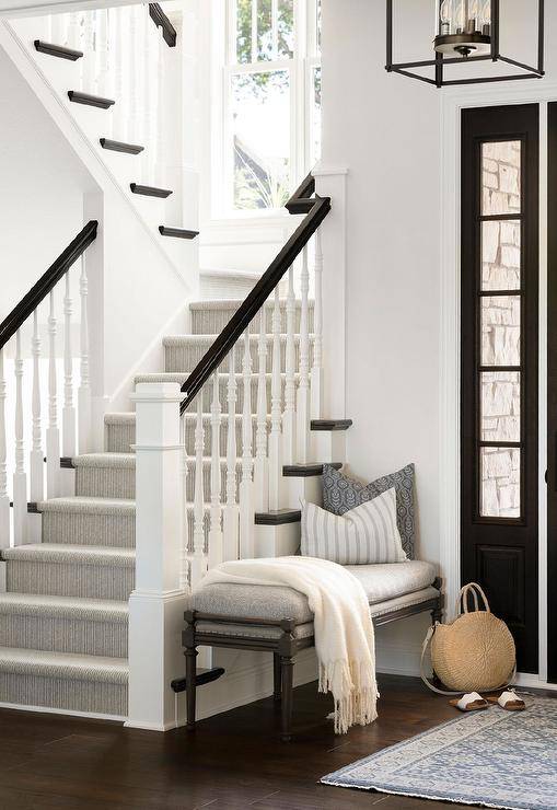 A white and gray rug sits in front of a black front door and a gray French bench positioned against a small staircase wall. The staircase is accented with a gray stripe stair runner covering black treads, while a black wooden handrail is fixed to white spindles.