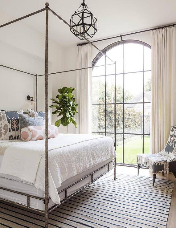 Bedroom features a silver canopy bed with pink and gray Ikat pillows illuminated by a Suzanne Kasler Morris lantern and a stripe rug.