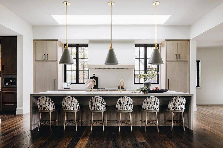 A row of black and white upholstered stools are placed in a brown kitchen at a light brown chevron island lit by gold and black conical pendants hung over a trough sink with
